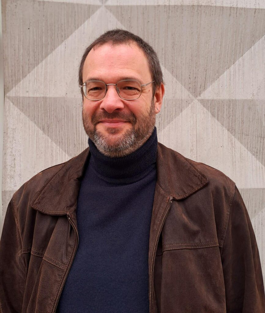 Mann mit Bart und Brille lächelt vor Wand.