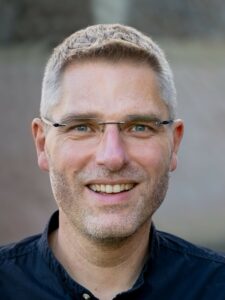 Mann mit Brille und Bart lächelt draußen.