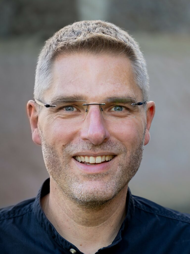 Mann mit Brille und Bart lächelt draußen.