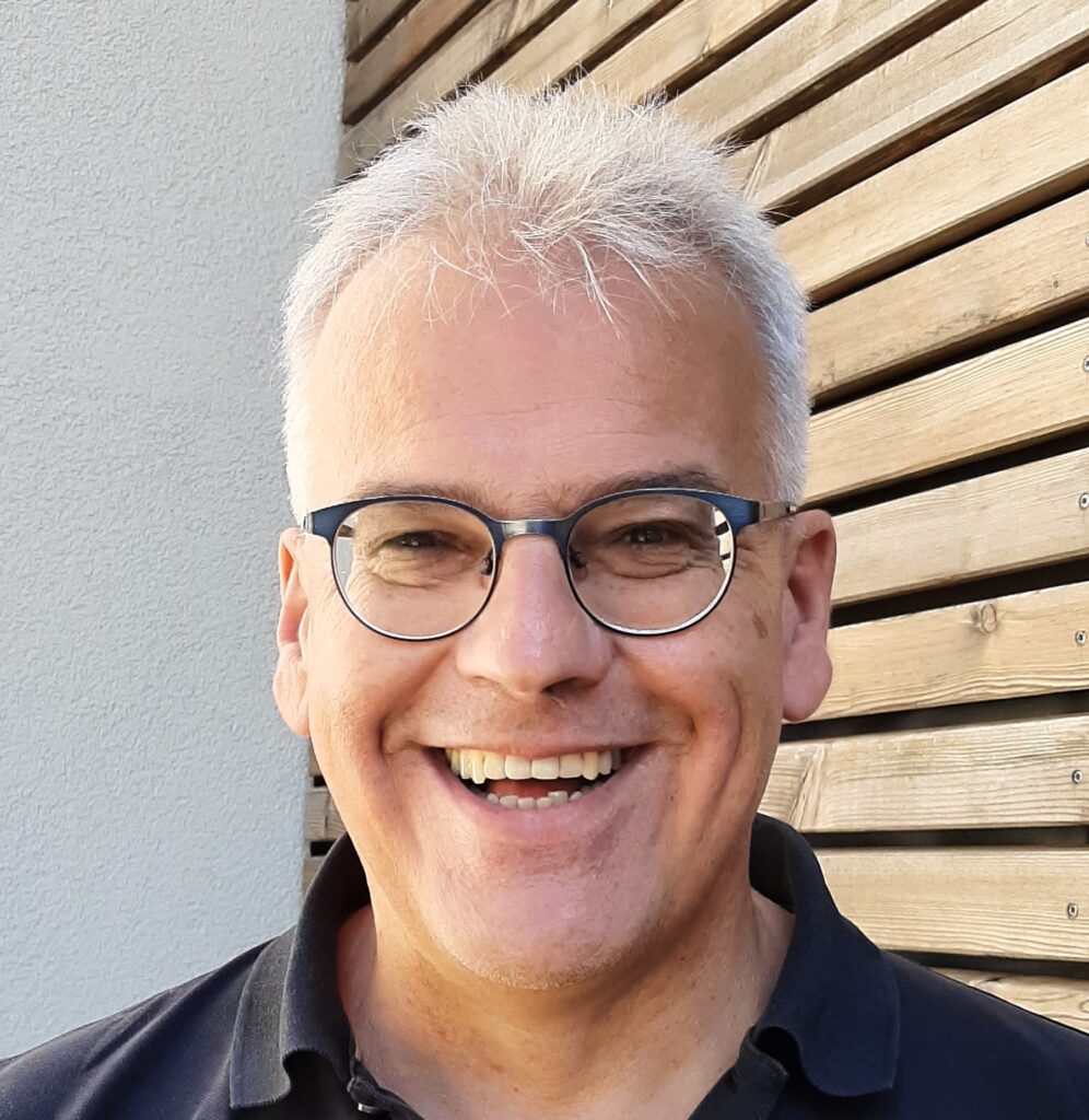 Lächelnder Mann mit Brille vor Holzachtergrund.