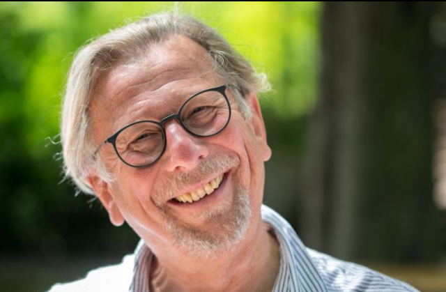 Ein älterer Mann mit Brille, grauen Haaren und einem kurzen Bart lächelt warm im Freien, wobei die grünen Bäume im Hintergrund verschwimmen. Er trägt ein gestreiftes Hemd und scheint einen sonnigen Tag zu genießen.