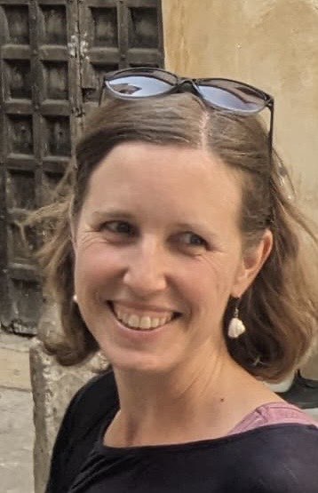 Eine Frau mit kurzen braunen Haaren lächelt, trägt eine Sonnenbrille auf dem Kopf, Ohrringe und ein schwarzes Oberteil und steht im Freien vor einer rustikalen Holztür.
