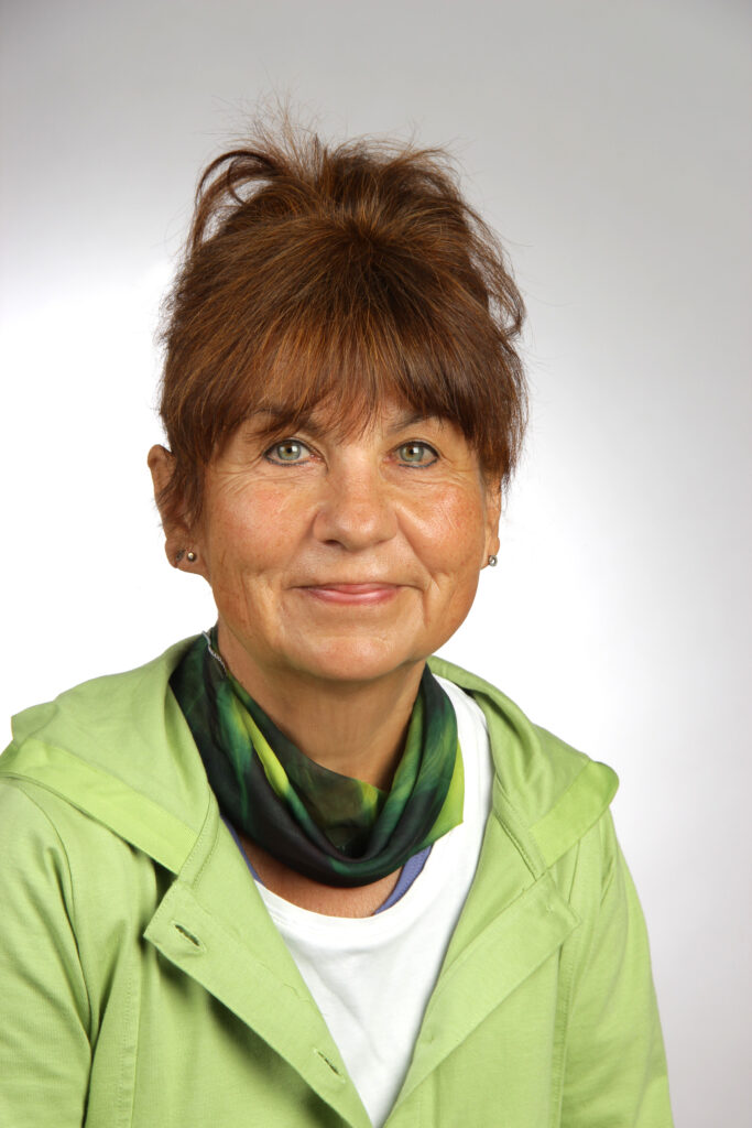 Eine Frau mit braunem Haar, die eine hellgrüne Jacke, einen dunkelgrünen Schal und ein weißes Oberteil trägt, lächelt in die Kamera vor einem schlichten hellen Hintergrund.