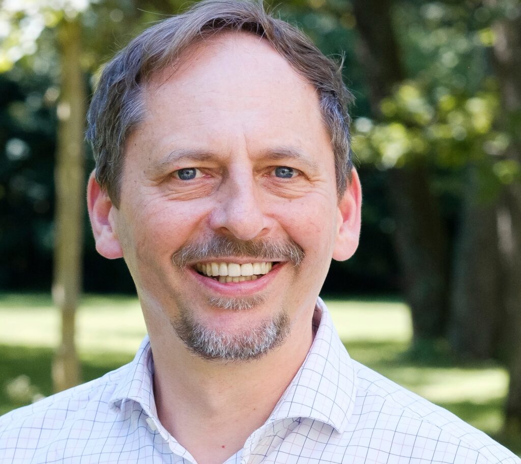 Ein Mann mit hellbraunem Haar und Ziegenbart lächelt im Freien, er trägt ein helles, kariertes Hemd, im Hintergrund sind Bäume und Grünpflanzen zu sehen.