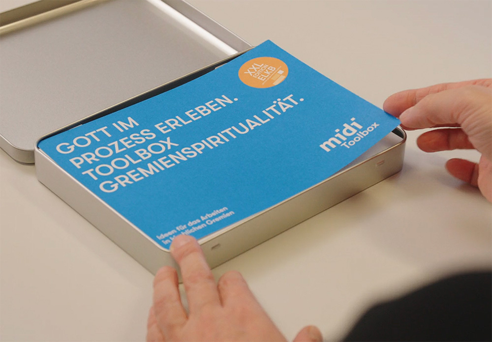 Eine Person öffnet eine Metallbox, die ein blaues Heft mit dem Titel GOTT IM PROZESS ERLEBEN: TOOLBOX GREMIENSPIRITUALITÄT von midi toolbox auf einer weißen Fläche enthält.