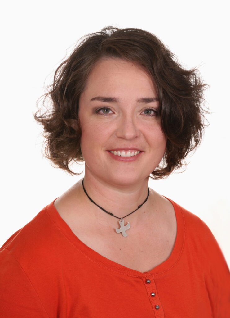 Frau mit kurzen, gewellten braunen Haaren lächelt in die Kamera. Sie trägt ein orangefarbenes Oberteil und eine Halskette mit einem silbernen Anhänger vor einem schlichten weißen Hintergrund.