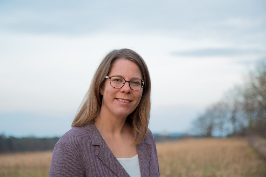 Eine Frau mit schulterlangem blondem Haar und Brille, die einen lilafarbenen Blazer trägt, steht im Freien auf einem Feld mit Bäumen und einem bewölkten Himmel im Hintergrund.