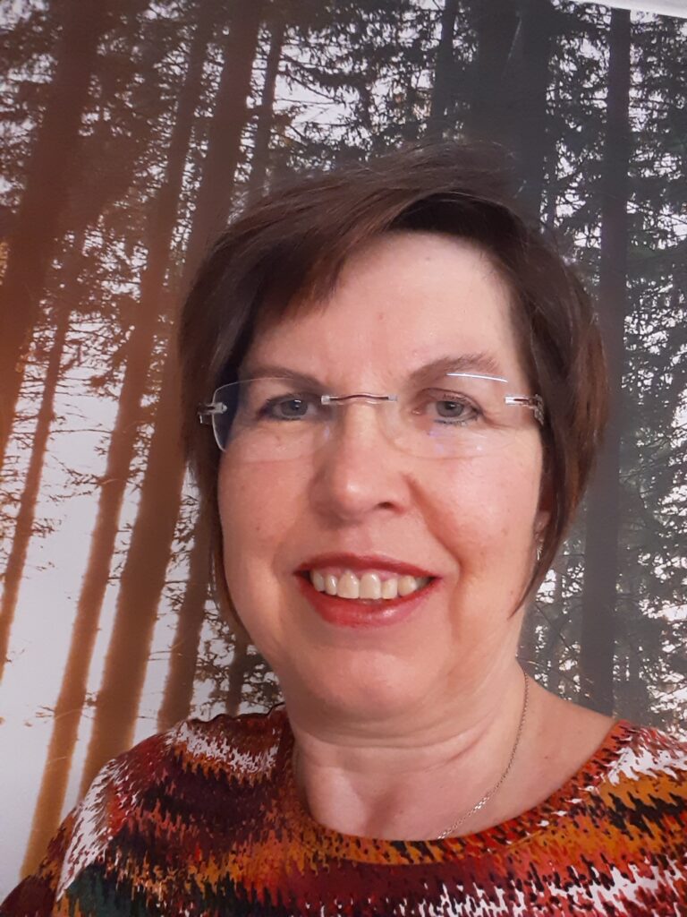 Eine Frau mit kurzen braunen Haaren und Brille lächelt in die Kamera und steht vor einem waldähnlichen Hintergrund mit hohen Bäumen.