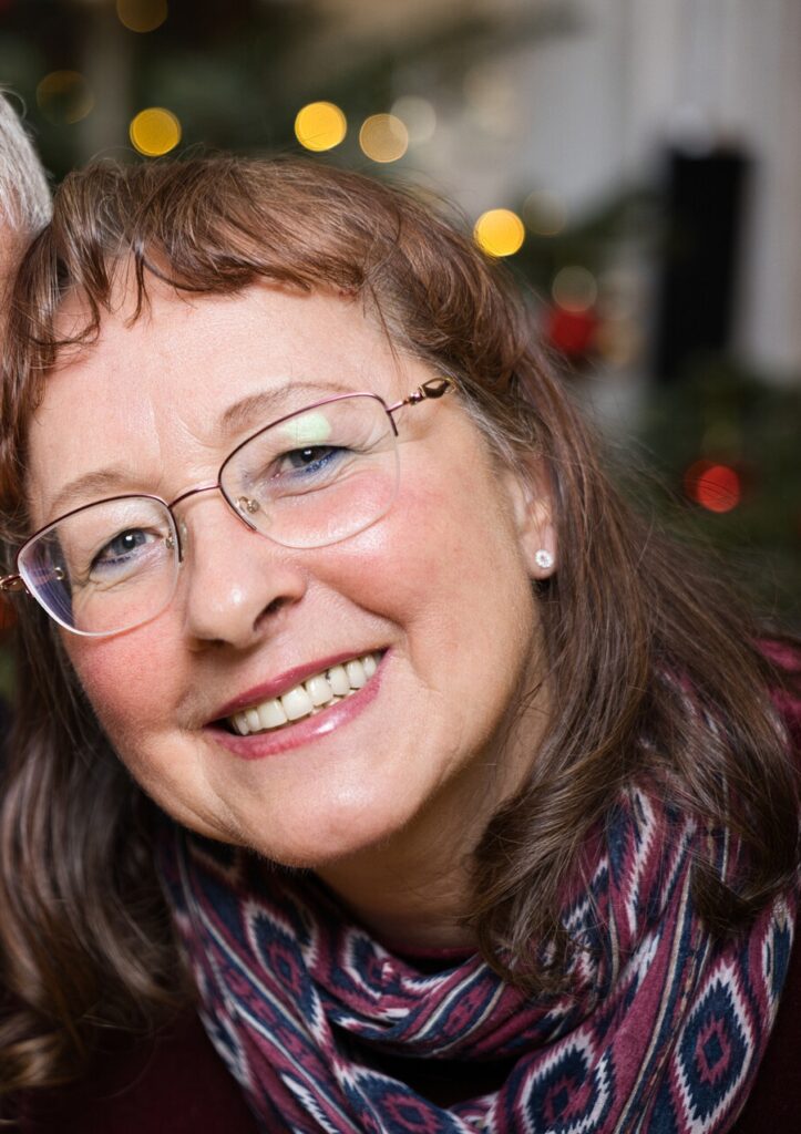 Eine lächelnde Frau mit Brille und braunem Haar trägt einen gemusterten Schal und posiert in einem Innenraum mit verschwommenen festlichen Lichtern im Hintergrund.