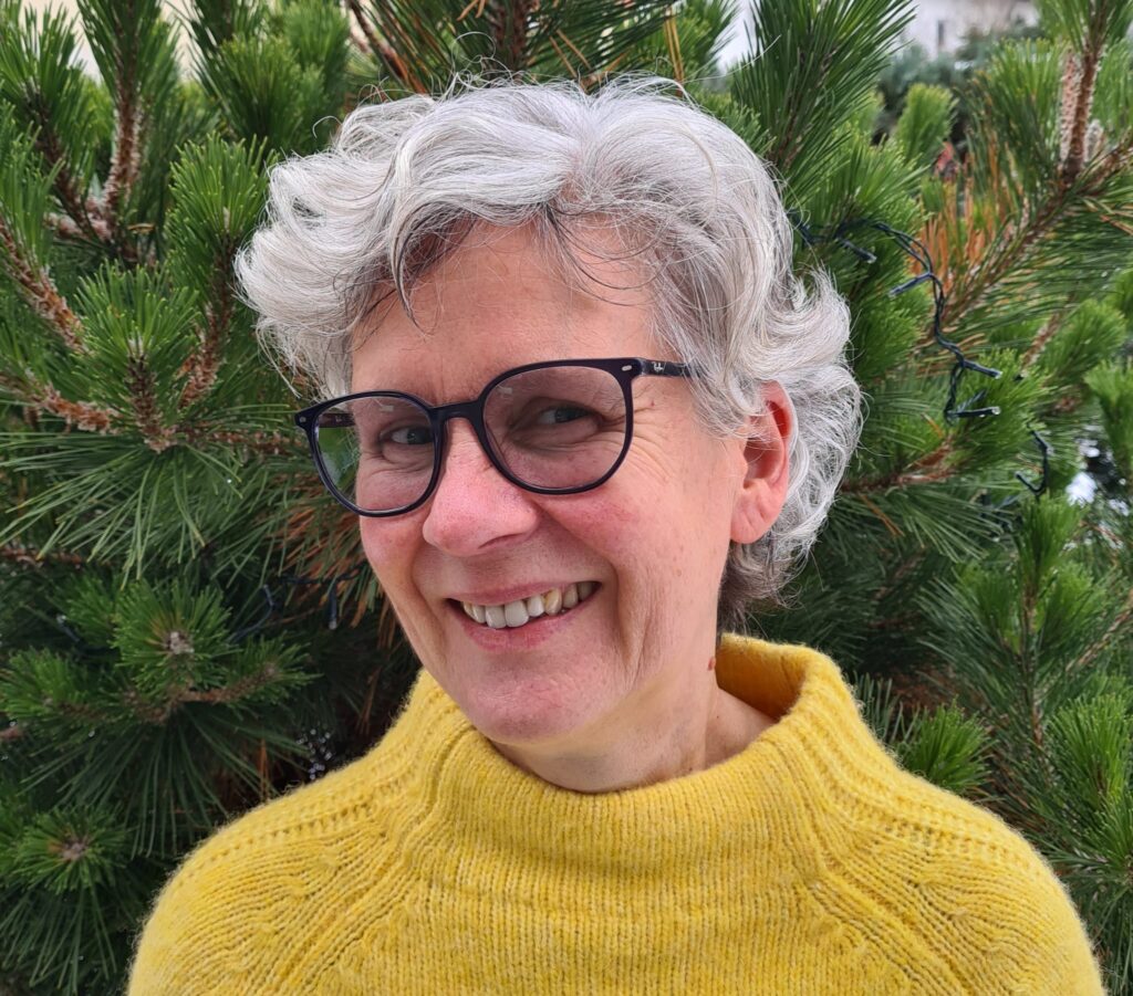 Lächelnde ältere Frau mit kurzen grauen Haaren und Brille, die einen gelben Pullover trägt, steht im Freien vor grünen Kiefern.
