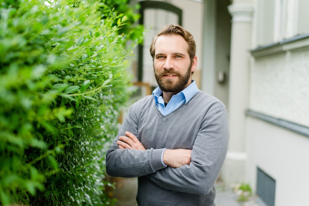 Ein Mann mit Bart und Schnurrbart, der einen grauen Pullover über einem blauen Hemd trägt, steht draußen mit verschränkten Armen neben einer grünen Hecke.
