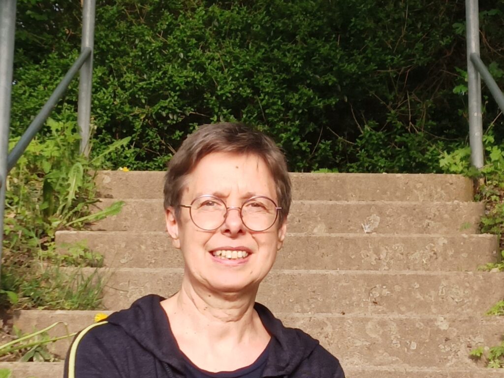 Eine Frau mit kurzen Haaren und Brille sitzt an einem sonnigen Tag lächelnd im Freien vor einer Steintreppe, die von Grün umgeben ist.