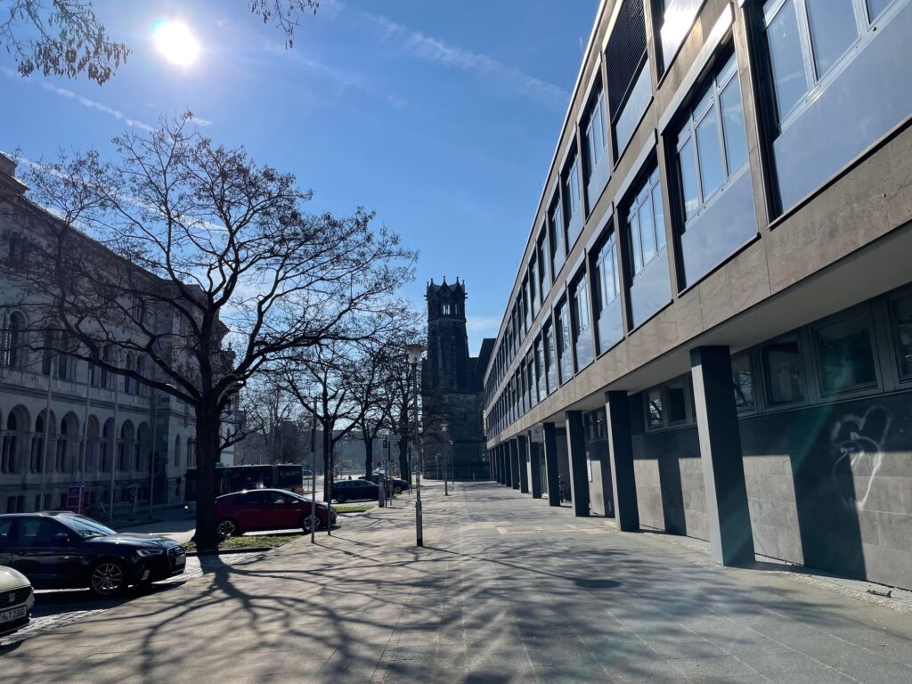 Sonnige Straßenszene mit blattlosen Bäumen, geparkten Autos, einem modernen Glasgebäude auf der rechten Seite und einem historischen Uhrenturm im Hintergrund unter blauem Himmel.