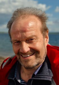 Ein Mann mittleren Alters mit hellbraunem Haar und Bart lächelt im Freien. Er trägt eine rote Jacke und steht an einem Gewässer mit einem bewölkten blauen Himmel im Hintergrund.