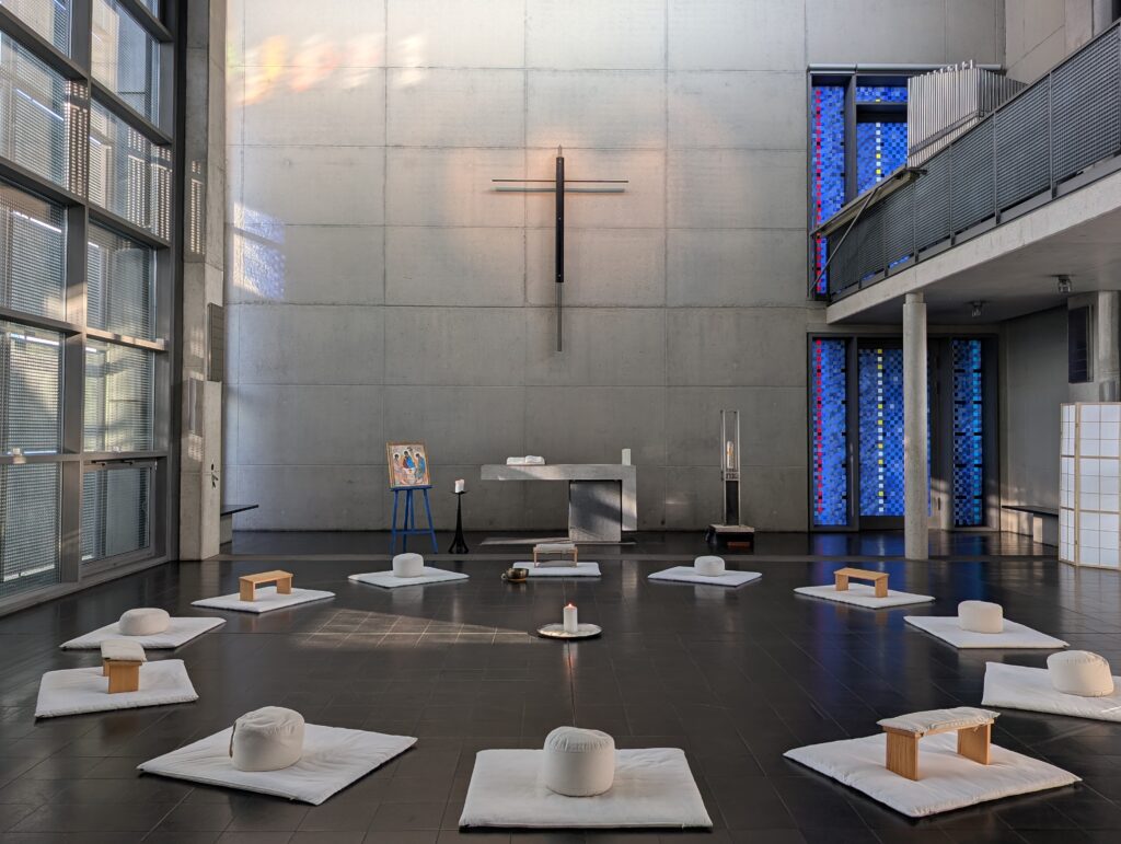 Eine moderne Kapelle mit einem Kreuz an einer Betonwand, einem Altar und weißen Meditationskissen, die im Kreis auf dem Boden angeordnet sind. Rechts befinden sich blaue Buntglasfenster.