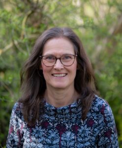 Eine Frau mit Brille und langen braunen Haaren lächelt im Freien und trägt ein blau gemustertes Oberteil mit roten und weißen Blumenmustern. Grünes Laub ist unscharf im Hintergrund zu erkennen.