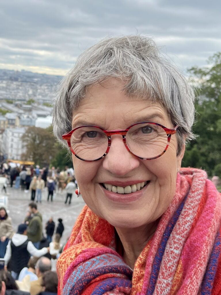 Lächelnde ältere Frau mit kurzen grauen Haaren und roter Brille, die einen bunten Schal trägt, im Freien mit einer Stadtlandschaft und einer Menschenmenge im Hintergrund.