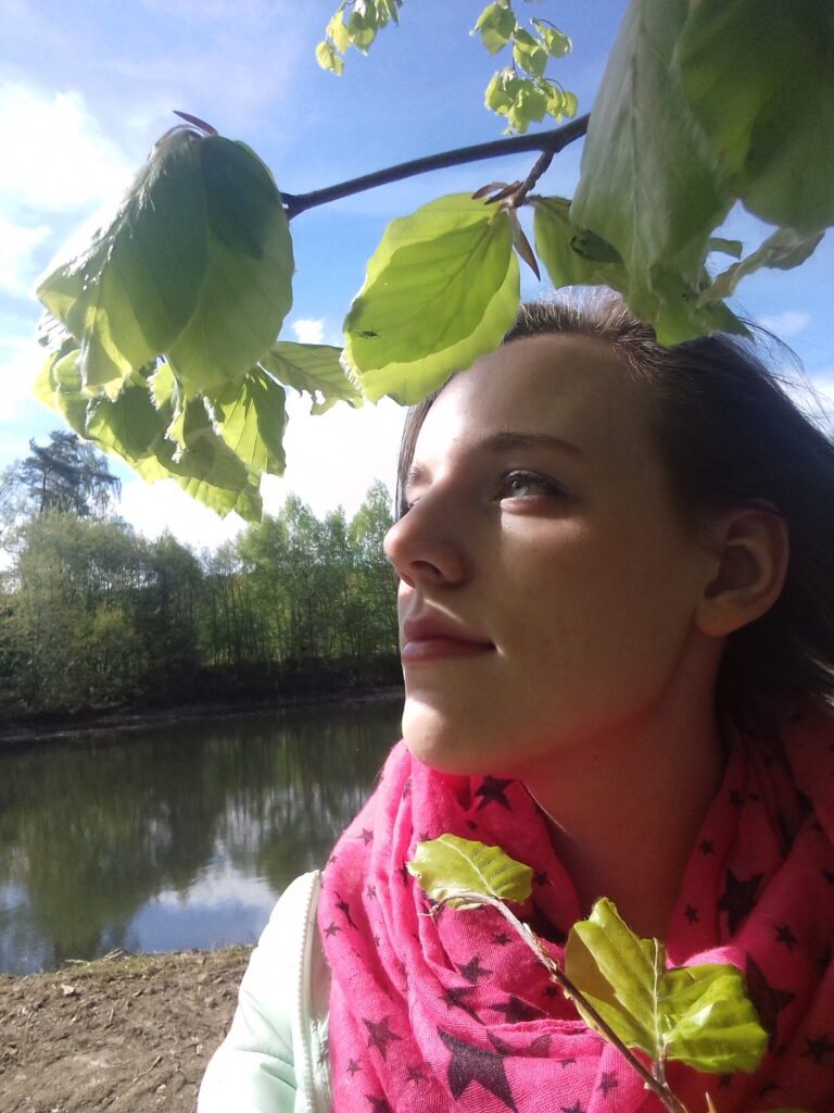 Eine Person mit einem rosafarbenen, mit Sternen gemusterten Schal steht an einem Fluss und blickt auf grüne Blätter über dem Kopf, mit Bäumen und blauem Himmel im Hintergrund.
