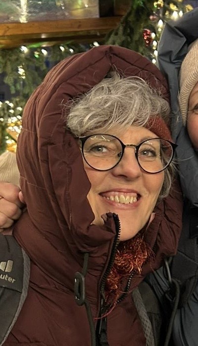 Eine lächelnde Person mit grauem Haar und Brille, die eine kastanienbraune Kapuzenjacke trägt, steht im Freien vor festlichen Lichtern und Grünpflanzen im Hintergrund.
