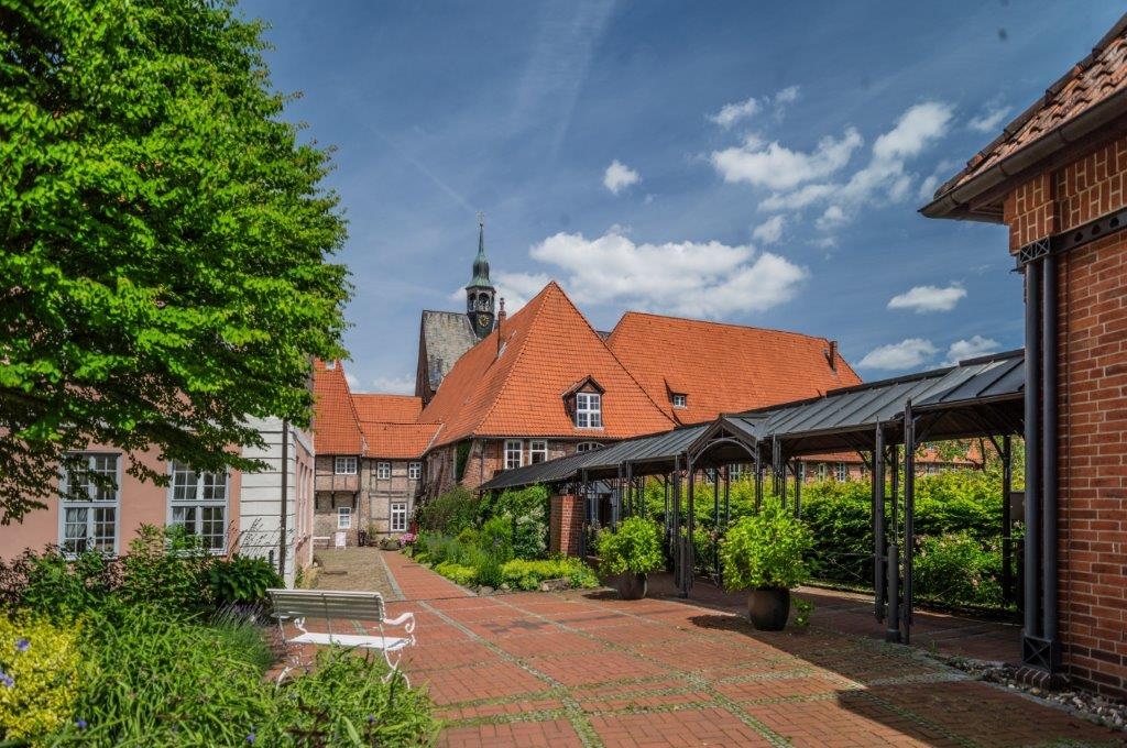 Ein Hof mit roten Ziegelgebäuden, orangefarbenen Ziegeldächern, grünen Bäumen, Topfpflanzen und einem Kirchturm im Hintergrund unter einem blauen Himmel mit Wolken.