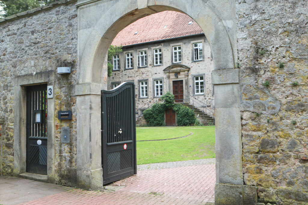 Ein steinernes Tor öffnet sich zu einem Innenhof mit einem historischen Gebäude im Hintergrund; das Gebäude ist mit Efeu bewachsen und eine Treppe führt zum Eingang.