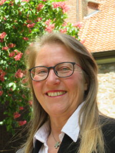 Eine lächelnde Frau mit Brille, die einen schwarzen Blazer und ein weißes Hemd trägt, steht im Freien in der Nähe von blühenden rosa Blumen und einem Gebäude mit Ziegeldach.