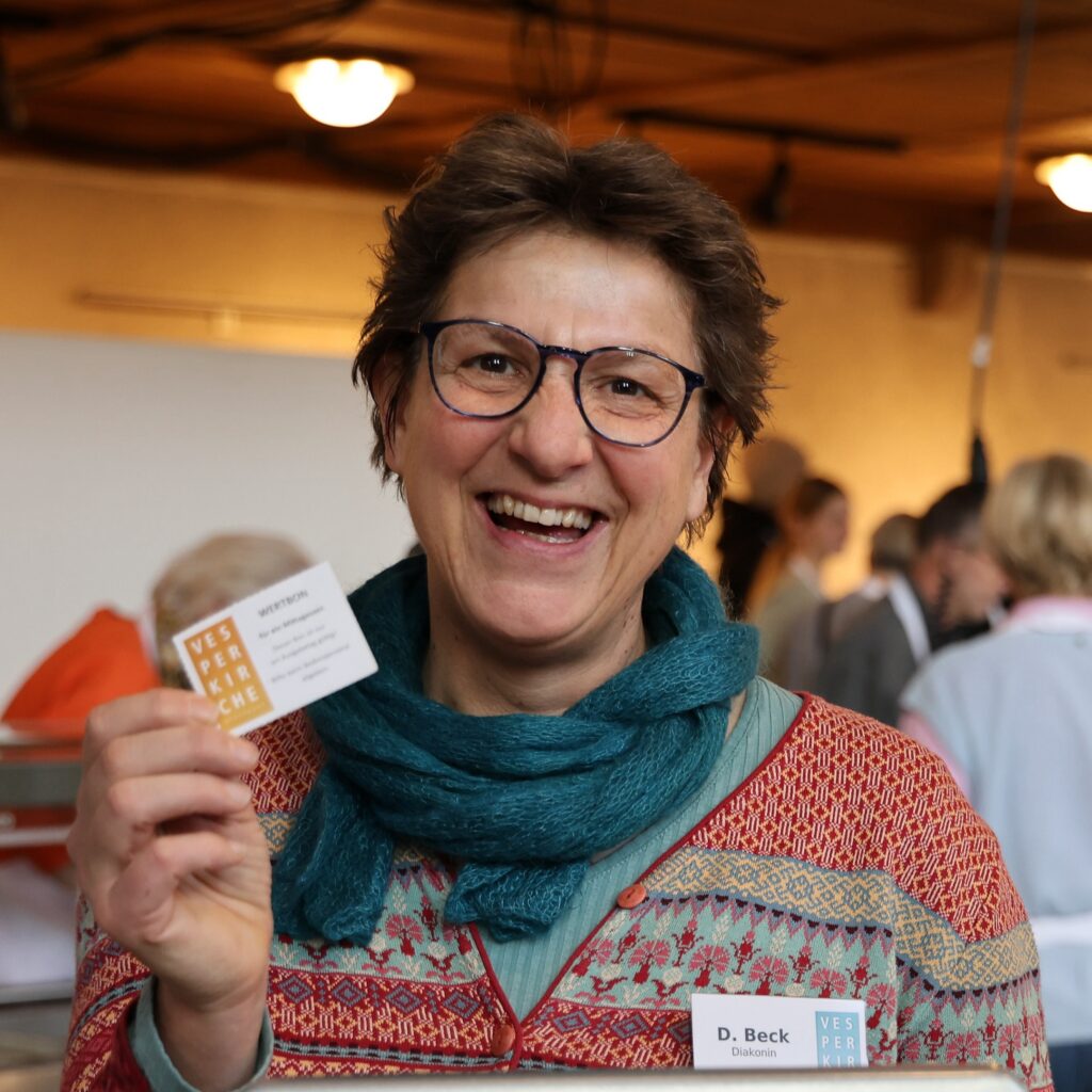 Eine lächelnde Person mit kurzen braunen Haaren und Brille hält eine Visitenkarte hoch. Sie trägt einen gemusterten Pullover und einen blaugrünen Schal und steht in einem Haus mit anderen Personen im Hintergrund.