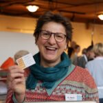 Lächelnde Person mit kurzen Haaren und Brille hält eine Visitenkarte hoch; trägt ein gemustertes Oberteil, einen blaugrünen Schal und ein Namensschild mit der Aufschrift D. Beck. Innenbereich mit anderen Personen unscharf.
