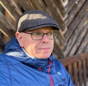 Ein Mann mit Brille und flacher Mütze, der eine blaue Jacke trägt, sitzt im Freien vor einer Holzwand und blickt im natürlichen Licht nachdenklich zur Seite.