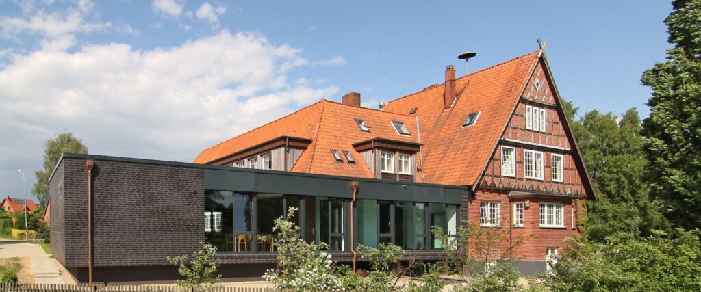 Ein modernes Gebäude aus Glas und Backstein ist an ein traditionelles Haus mit rotem Dach und hölzernen Details angebaut, umgeben von Grün und einem teilweise bewölkten Himmel.