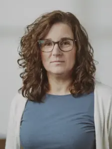 Eine Frau mit lockigem braunem Haar und Brille, die ein blaues Oberteil und eine helle Strickjacke trägt, blickt vor einem neutralen Hintergrund ruhig in die Kamera.