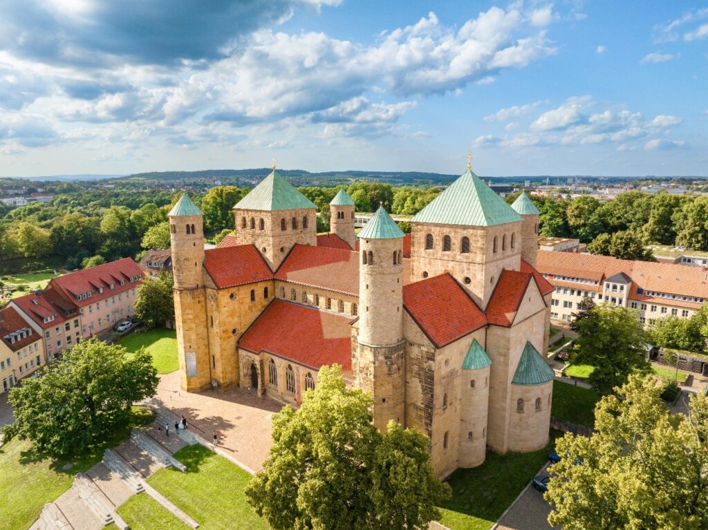 Luftaufnahme einer großen romanischen Kirche mit vier Türmen, rotem Ziegeldach und grünen Turmspitzen, umgeben von Grünflächen und nahe gelegenen Gebäuden unter einem teilweise bewölkten Himmel.