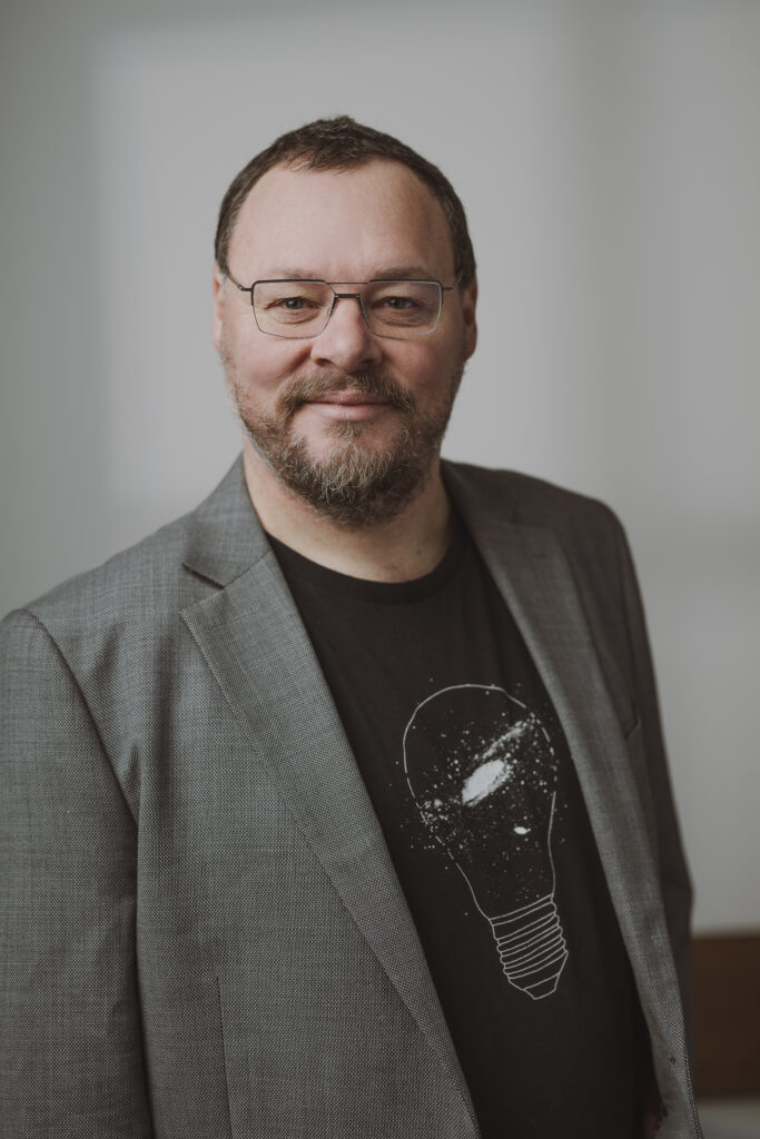 Ein Mann mit Brille und Bart, der einen grauen Blazer und ein schwarzes T-Shirt mit einer weißen Glühbirnen-Grafik trägt, steht lächelnd vor einem neutralen Hintergrund.