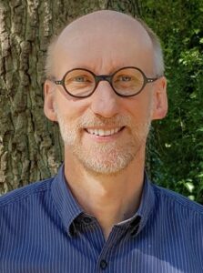 Ein lächelnder Mann mittleren Alters mit Brille und kurzem Bart steht im Freien vor einem Baum und trägt ein blaues Button-up-Hemd.