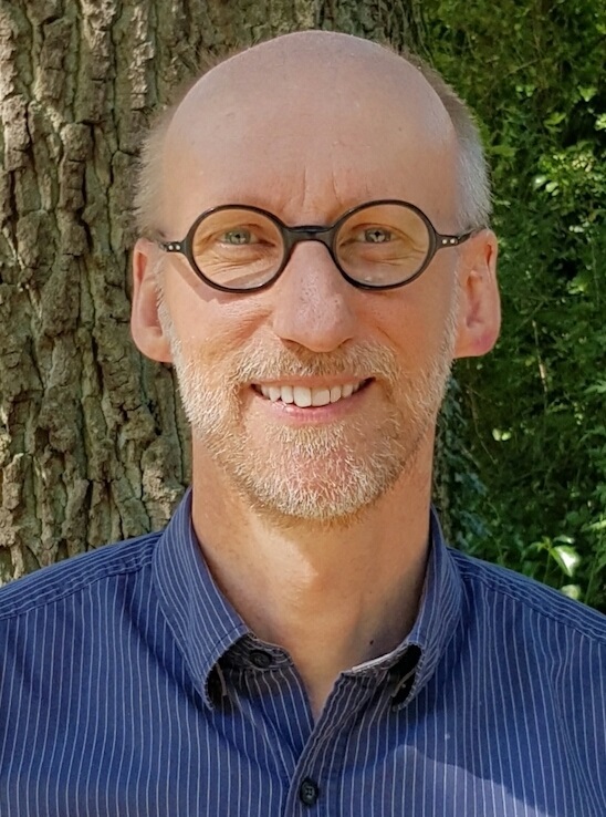 Ein lächelnder Mann mittleren Alters mit Brille und kurzem Bart steht im Freien vor einem Baum und trägt ein blaues Button-up-Hemd.