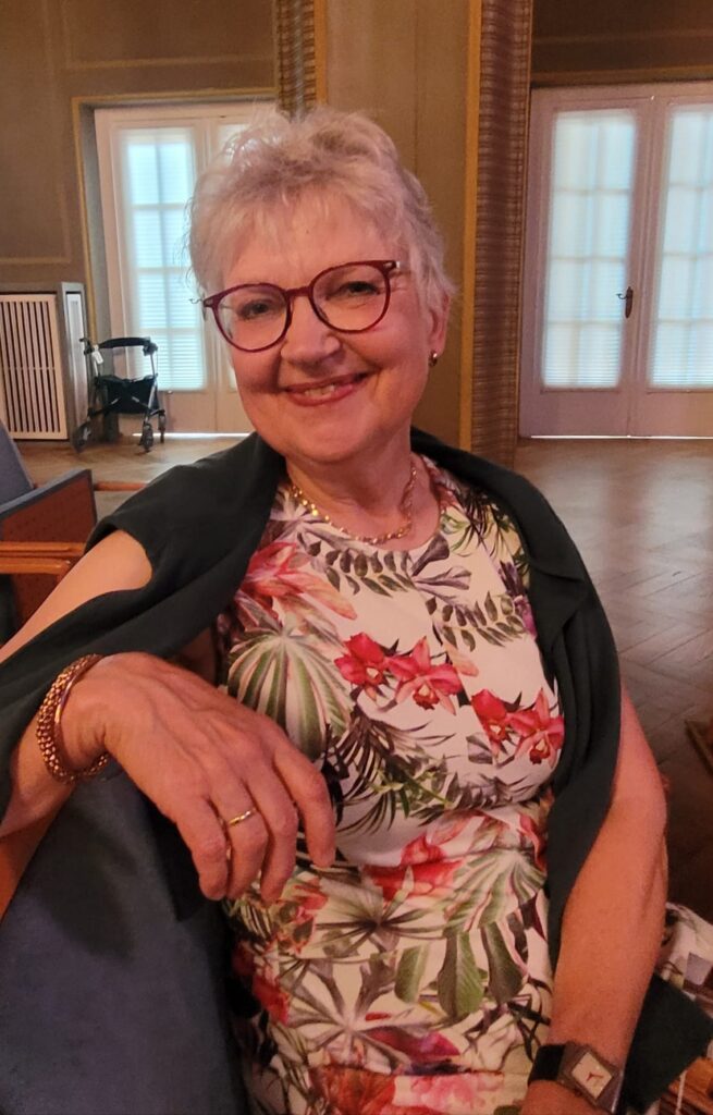 Lächelnde ältere Frau mit kurzen grauen Haaren und Brille, die ein geblümtes Kleid trägt und mit einem grünen Schal über den Schultern in einem Haus sitzt.