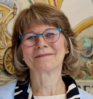 Eine lächelnde ältere Frau mit kurzen grauen Haaren und blauer Brille, die eine hellblaue Jacke und einen gemusterten Schal trägt, steht vor einem bunt bemalten Wandgemälde.