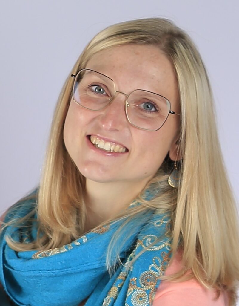 Eine lächelnde Frau mit langen blonden Haaren, die eine Brille und einen blau gemusterten Schal trägt, posiert vor einem hellen Hintergrund.