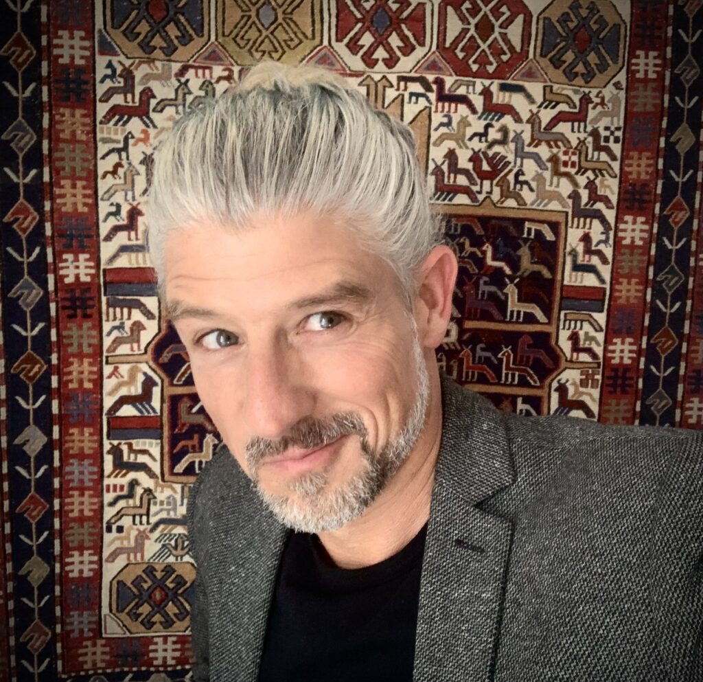 Ein Mann mit grauem Haar in einem Dutt und einem grauen Bart lächelt, während er einen grauen Blazer trägt und vor einem bunt gemusterten Teppich steht.