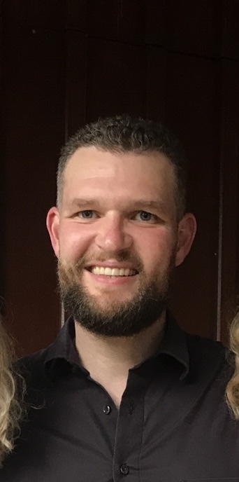 Ein Mann mit kurzem lockigem Haar und Bart, der ein schwarzes Hemd mit Kragen trägt, lächelt vor einem dunklen Holzhintergrund.