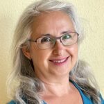 Eine lächelnde ältere Frau mit langen grauen Haaren, Brille und einem blaugrünen Oberteil steht vor einer hellgelben Wand.