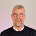 Ein lächelnder Mann mit kurzen blonden Haaren, Bart und Brille, der einen schwarzen Pullover über einem weißen Hemd trägt, steht vor einem hellen Hintergrund.