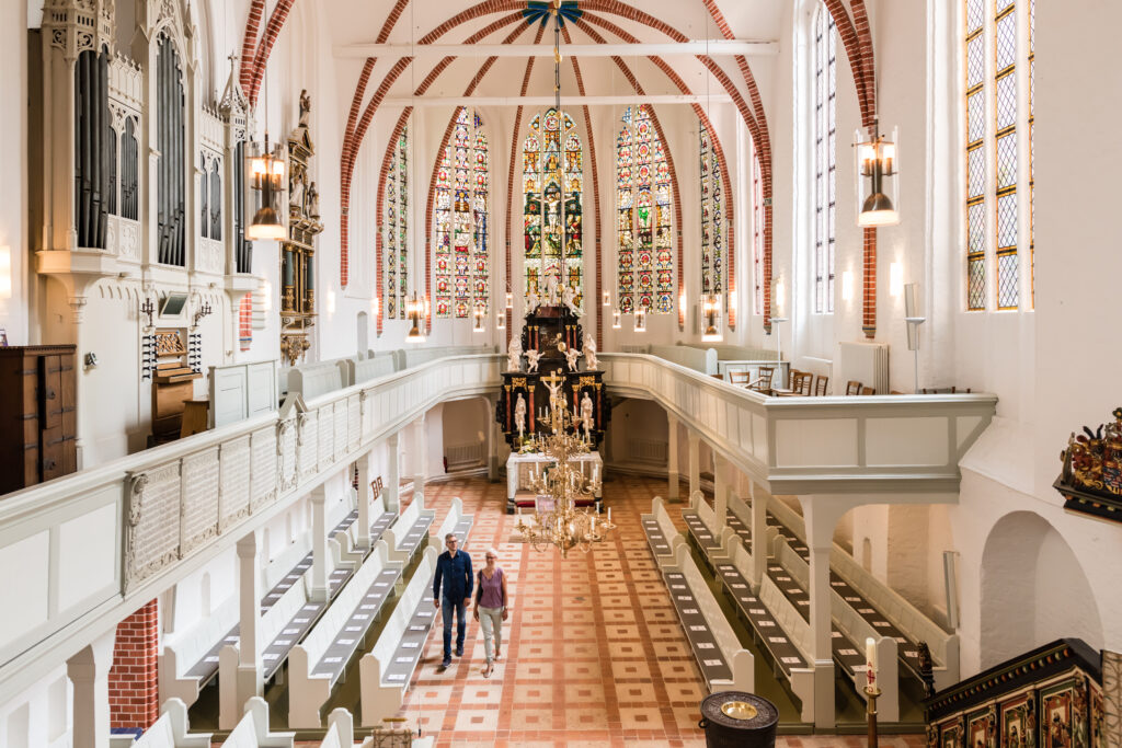 Das Innere einer hellen Kirche mit Buntglasfenstern, weißen Kirchenbänken, Kronleuchtern und zwei Personen, die den Mittelgang entlanggehen.