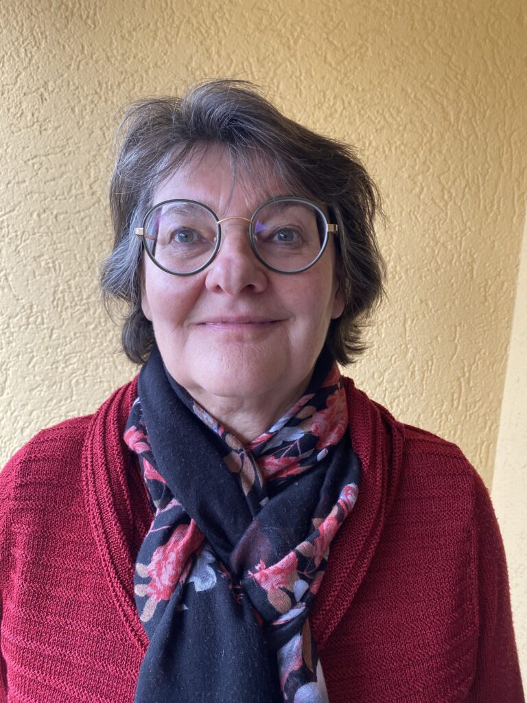 Frau mit kurzen grauen Haaren und Brille, die einen roten Pullover und einen geblümten Schal trägt, steht vor einer strukturierten gelben Wand und lächelt in die Kamera.