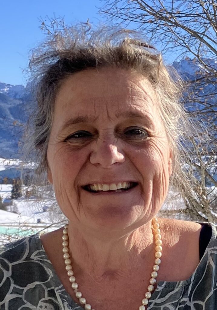 Lächelnde ältere Frau mit grauem Haar, die ein gemustertes Hemd und eine Perlenkette trägt, steht im Freien mit verschneiten Bergen und kahlen Bäumen im Hintergrund.