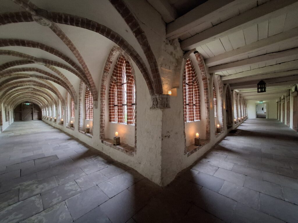 Ein steinerner Gang mit Gewölbedecken und mit Ziegeln eingefassten Fenstern, der an beiden Seiten von Kerzen beleuchtet wird und eine warme, historische Atmosphäre schafft.