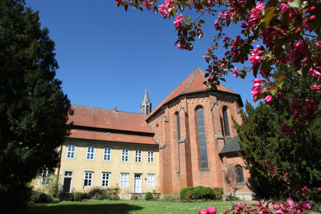 Neben einem gelben Gebäude steht eine historische Backsteinkirche mit hohen Bogenfenstern, umrahmt von blühenden rosa Blumen und grünen Bäumen unter einem strahlend blauen Himmel.