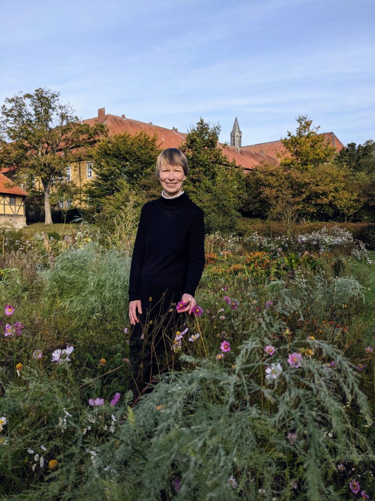 Eine Frau in einem schwarzen Pullover steht lächelnd in einem Wildblumengarten, im Hintergrund sind Bäume und historische Gebäude zu sehen, über ihr erstreckt sich ein klarer Himmel.