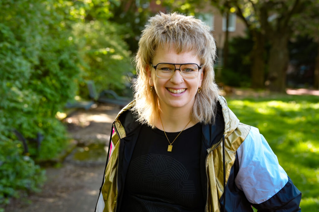 Eine lächelnde Person mit kurzen, gestuften blonden Haaren und Brille steht draußen in einem sonnendurchfluteten, grünen Park und trägt ein schwarzes Shirt und eine leichte Jacke.
