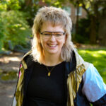 Eine lächelnde Person mit kurzen, strubbeligen blonden Haaren und Brille steht draußen in einem sonnendurchfluteten, grünen Park und trägt ein schwarzes Hemd und eine goldene Jacke.